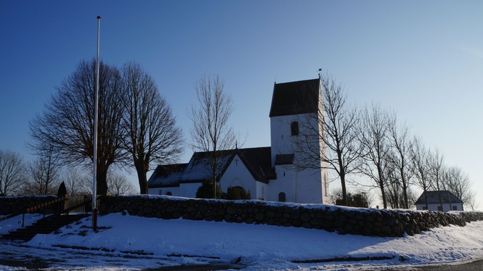 En hvid kirke med tårn og et mindre sidebygning, omgivet af sne og nakke træer.