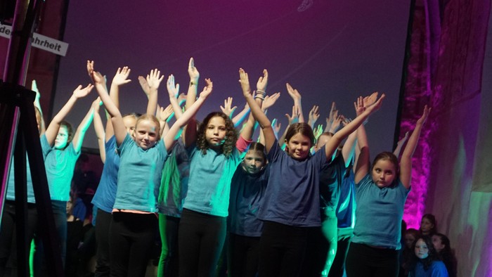 Eine Gruppe von Kindern hebt fröhlich die Arme auf der Bühne während einer Aufführung mit farbenfroher Beleuchtung.