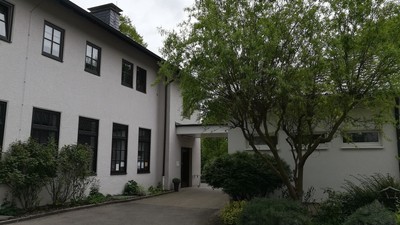 Das Bild zeigt ein weißes Gebäude mit schwarzen Fensterläden und einem Torbogen, umgeben von Bäumen und Büschen.