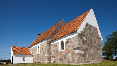 En lille kirke med røde tag og murstensmurer, placeret på en grøn eng med klar blå himmel.