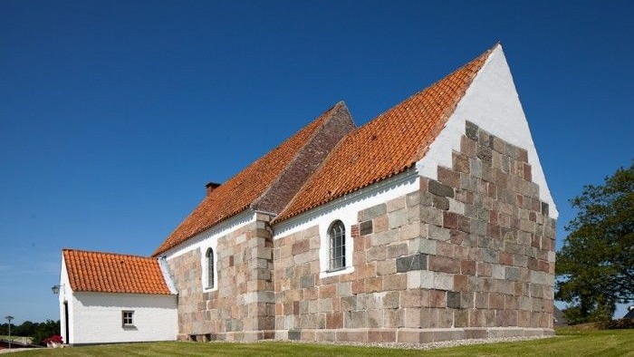 En lille kirke med røde tag og murstensmurer, placeret på en grøn eng med klar blå himmel.