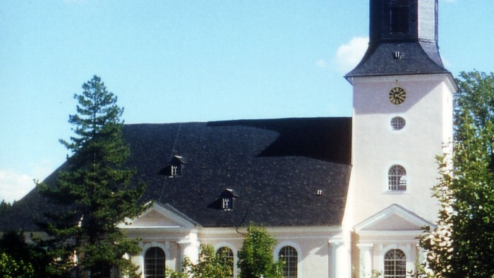 Elegante weiße Kirche mit hohem Glockenturm und Uhr, umgeben von üppigem Grün unter einem blauen Himmel.