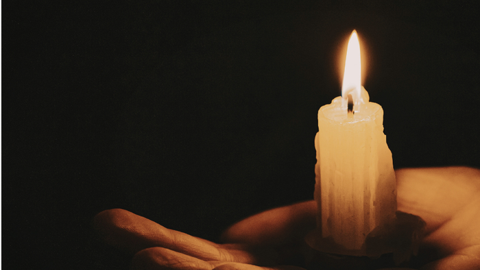 Et blødt oplyst stearinlys, der holdes varsomt mellem to hænder i et mørkt miljø.