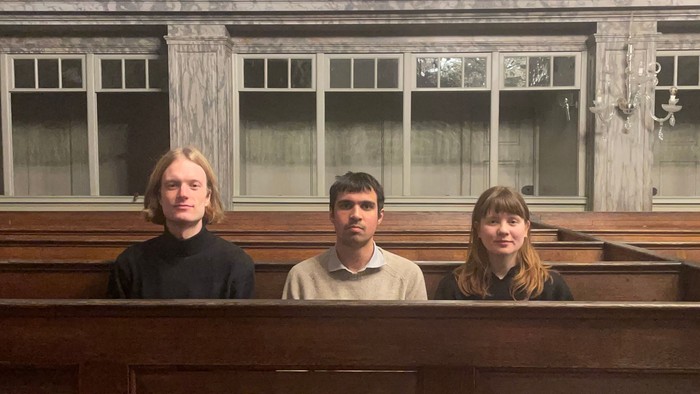 Tre personer poser sammen på en gammel trækirkebænk foran en klassisk interiørbaggrund.