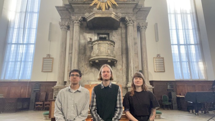 Tre personer sidder på kirkebænke foran en prædikestol med udsmykning og farvede glasvinduer.