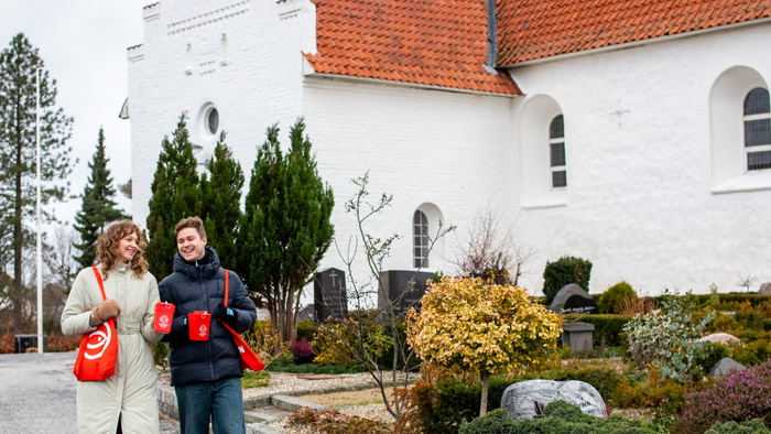 To mennesker går gennem et fredeligt haveområde nær en hvid kirke med rødt tag.