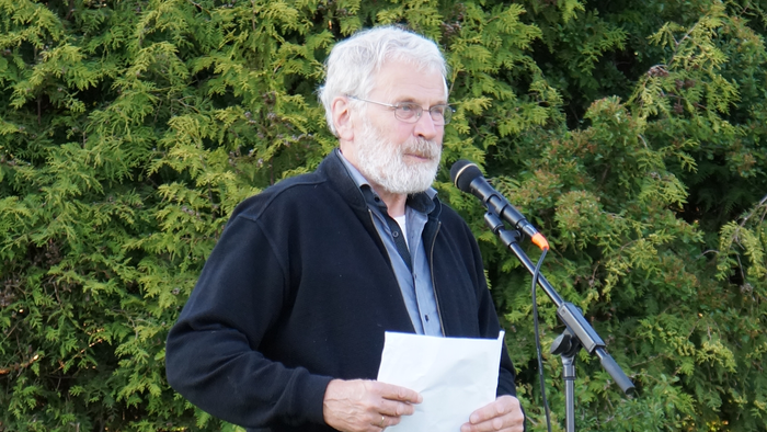 Mand med hvid skæg læser fra et papir ved et mikrofonstativ.