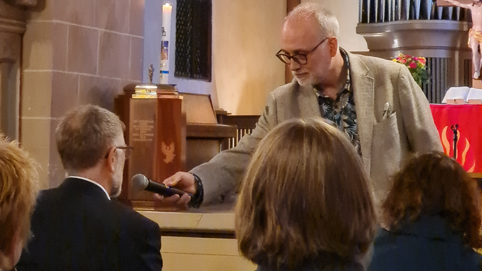 Ein Mann spricht während eines Gottesdienstes in ein Mikrofon, das ihm eine moderierende Person hinhält. Eine aufmerksame Gemeinde sitzt vor ihm.