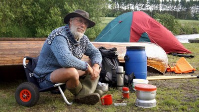 En mand sidder ved en vogn med campingudstyr og telte i en smuk udendørs omgivelse.