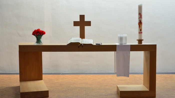 Minimalistischer Altar mit Kreuz, Blumen, Kerze und offenem Buch auf einem hölzernen Lesepult.