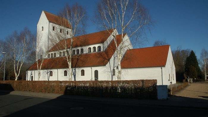 Et stort, hvidt og rødt taget kirkebygningskompleks med en høj tårn og flere sidebygninger, omgivet af nøgler og træer.