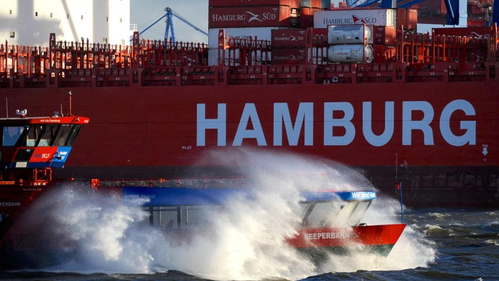 Ein großes Frachtschiff der Hamburg Süd navigiert, während ein Schlepper ihm in unruhigen Gewässern den Weg bahnt.