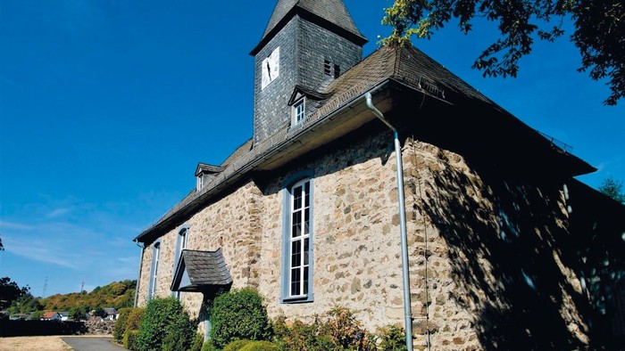Idyllische Steinkirche mit hohem Glockenturm und üppigem Grün drumherum.
