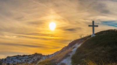 Ein friedlicher Sonnenuntergang über einem Hügel mit einem Kreuz, das sich gegen den leuchtenden Himmel abzeichnet.