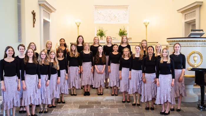 En gruppe kvinder i mørke tops og lyserøde skørter står sammen i et rum med marmorovne og lysekælder.