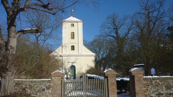 Weiße Kirche mit hohem Turm und grüner Tür, umgeben von Schnee und Bäumen.