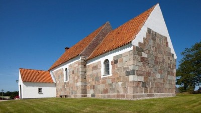 En lille kirke med røde tag og murstensmurer, placeret på en grøn eng med klar blå himmel.