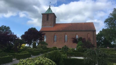 Hyggelig rødstenskirke med grønt tag og en velplejet havegang i fredfyldte omgivelser.