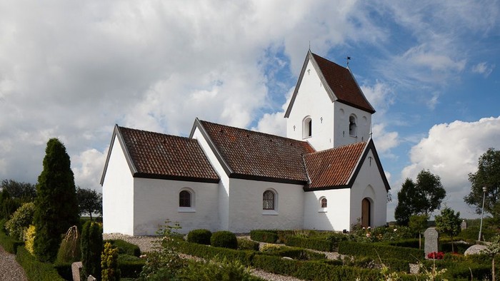 En hvid kirke med røde tag og tårn, omgivet af grønne buske og en klar blå himmel med hvidt skyer.