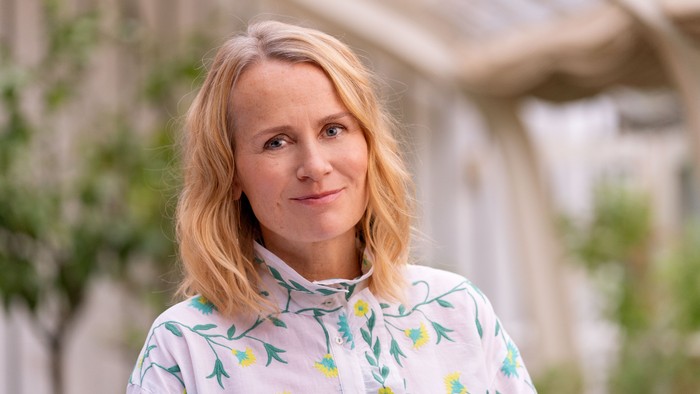 En kvinde med blondt hår smiler udendørs i en blomstret skjorte.