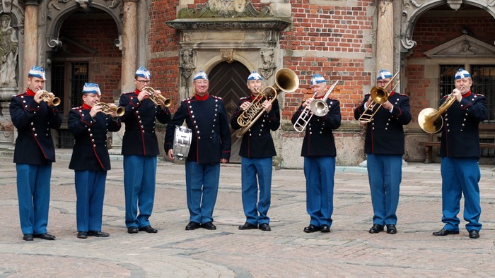 Musikanter i uniform med blå bukser og jakker spiller messingblæsere i en historisk gårdhave.