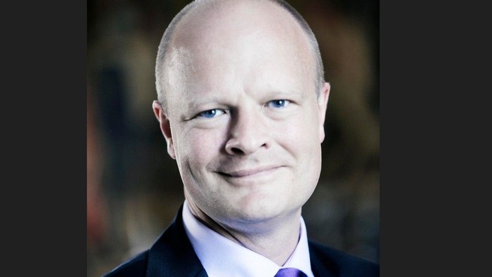 Professionelt portræt af en skaldet mand i mørkt jakkesæt, hvid skjorte og lilla slips, der smiler let.