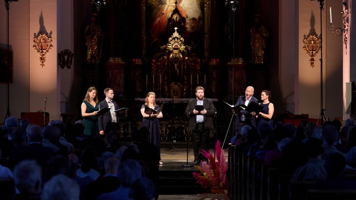 Et kvartet optræder på scenen i en rigt udsmykket kirke, oplyst af dramatisk belysning.