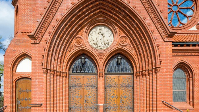 Gotischer Backsteinkircheneingang mit verzierten Holztüren, Bogenfenstern und einem kreisförmigen Buntglasrelief darüber.