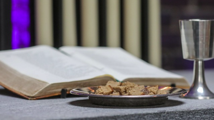 Offene Bibel neben Abendmahlsutensilien, darunter Brot und ein Kelch, auf einer dunklen Oberfläche.