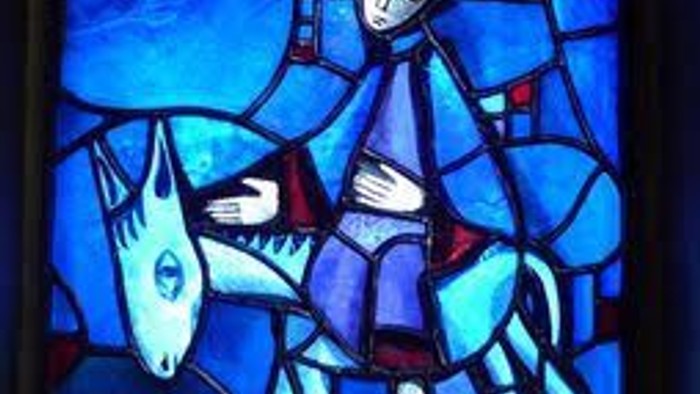 Bemalte Glasdarstellung einer in Roben gekleideten Figur, die auf einem Esel reitet, vor einem blauen Himmelshintergrund.