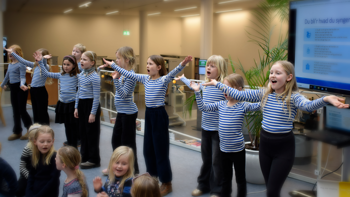 En gruppe børn danser og synger i en hall med stribede trøjer og mørke bukser.