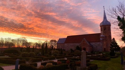 En kirke med tårn og klokke i solnedgang med rød og orange himmel, omgivet af en gravplads og grønne planter.