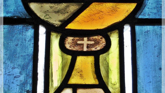 Das Bild zeigt ein buntes Kirchenfenster mit einem Kelch und Kreuz.