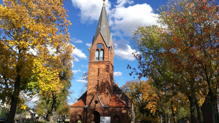 Kirchturm aus rotem Backstein, umgeben von Herbstbäumen unter blauem Himmel