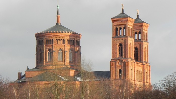 Zwei Türme einer Kirche mit spitzen Dächern und goldenen Akzenten, umgeben von Bäumen und einem bewölkten Himmel