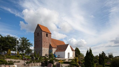 En lille kirke med tårn og røde tag i en trædiget have under et blødt blå himmel.
