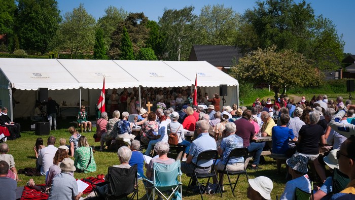 Udendørs arrangement med publikum siddende under et telt og overværer en sceneoptræden eller ceremoni på en solskinsdag.
