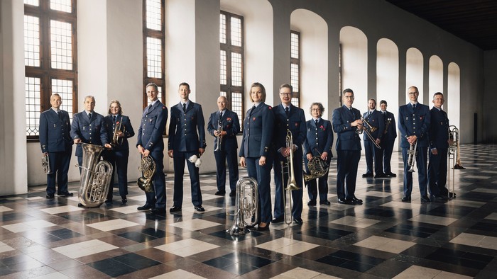 Uniformerede musikere poser formelt i en stor sal med klassiske instrumenter, stående i en ordnet række.