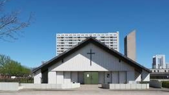 Moderne kirke med et kors på et simpelt gavltag, omgivet af bybygninger under en klar blå himmel.