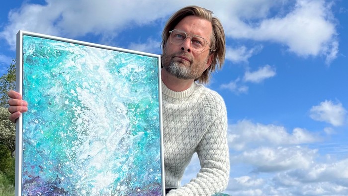 En mand knæler ved kysten og holder et abstrakt maleri af et kystlandskab, omgivet af tang.