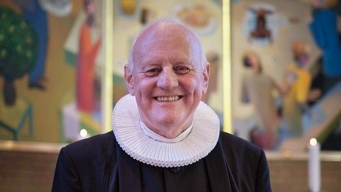 Smilende skaldet mand i gejstligt tøj med farverig muralbaggrund.