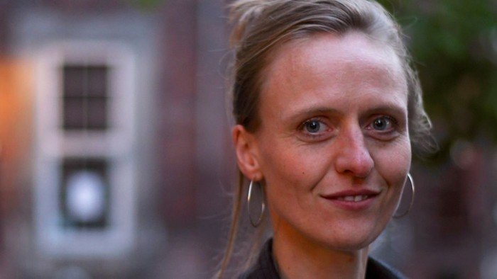 Kvinde med kort blondt hår og store øreringe, der smiler udendørs nær en bygning.