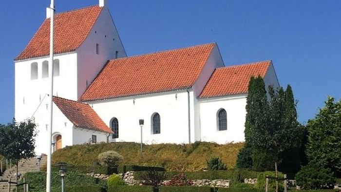 En charmerende hvid kirke med rødt tegltag placeret på en anlagt bakkeside under en klar blå himmel.