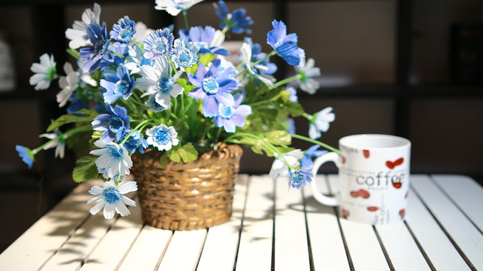 Ein charmanter Weidenkorb hält blaue und weiße Blumen neben einem gemütlichen Kaffeebecher auf einem Holztisch.