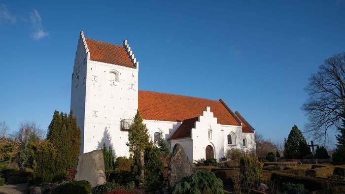 Et hvidt kirkebygning med røde tag og tårn, omgivet af grønne planter og træer under en klar blå himmel