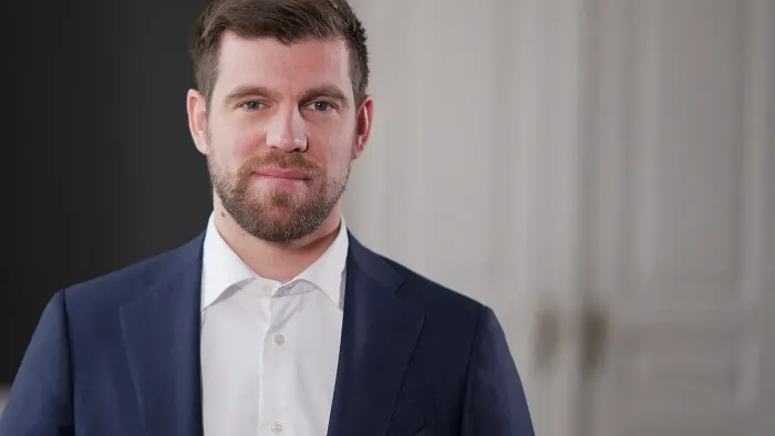 En mand i en mørk jakkesæt og hvid skjorte står selvsikkert foran en klassisk dørbaggrund.