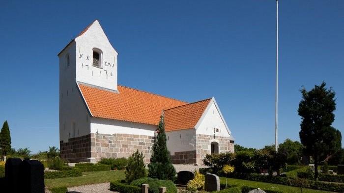 En hvid kirke med rød tag og tårn i en træket garden med gravsteder og et flagstang.
