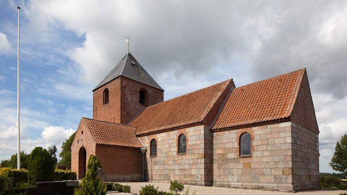 En lille kirke med tegltag og tårn i en trædit og rolig omgivelse