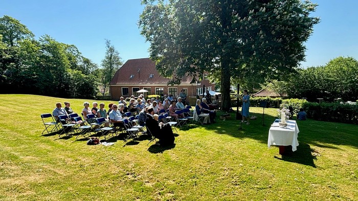En gruppe af mennesker sidder ude på græs og spiser, mens en person står ved en bord og taler.