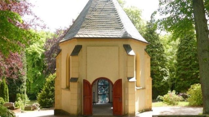 Ein kleines gelbes Gebäude mit spitzem Dach und zwei roten Türen in einem grünen Garten